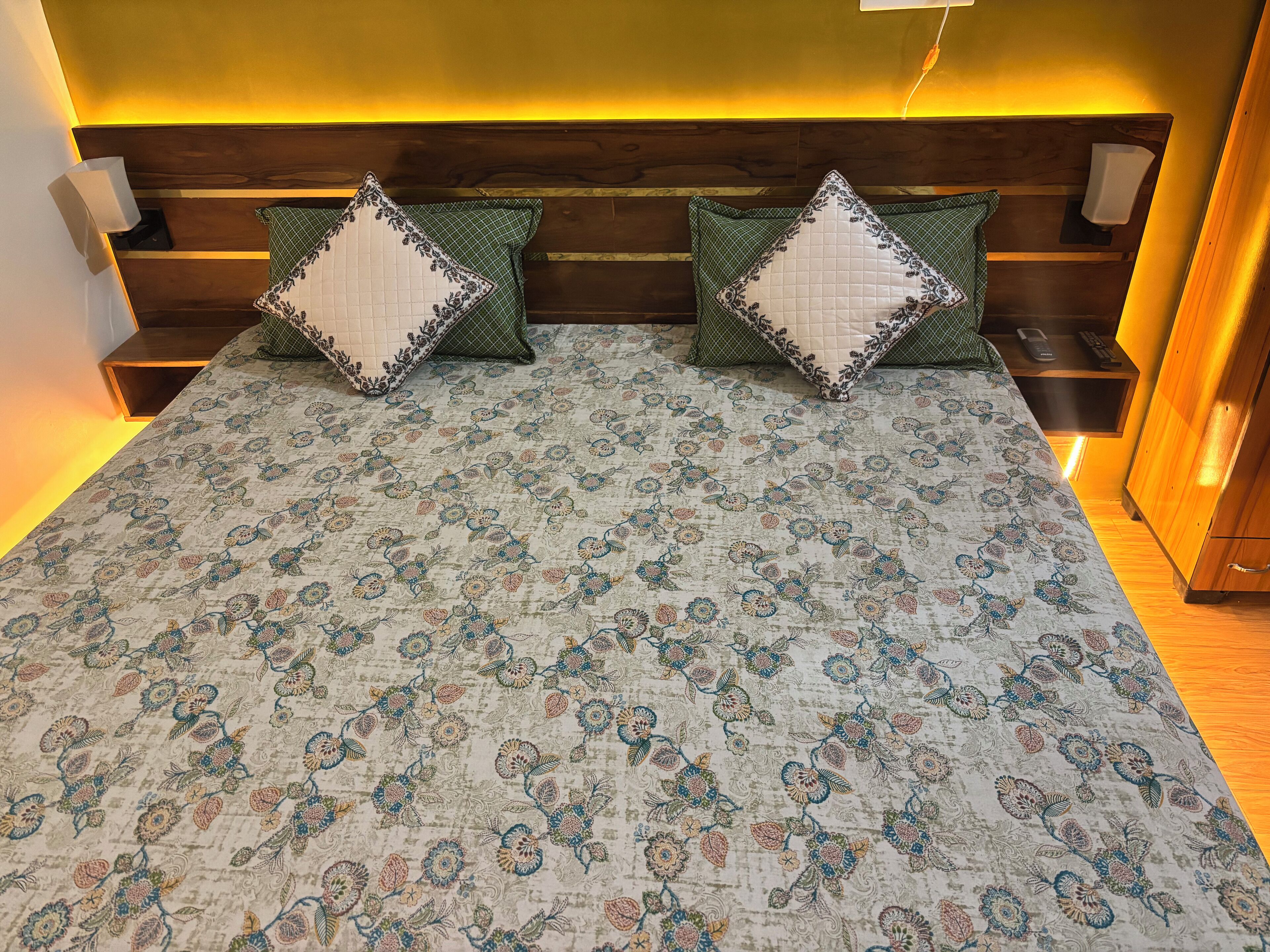 A bed with a floral-patterned bedspread, two decorative pillows, a wooden headboard, and bedside tables with lamps.