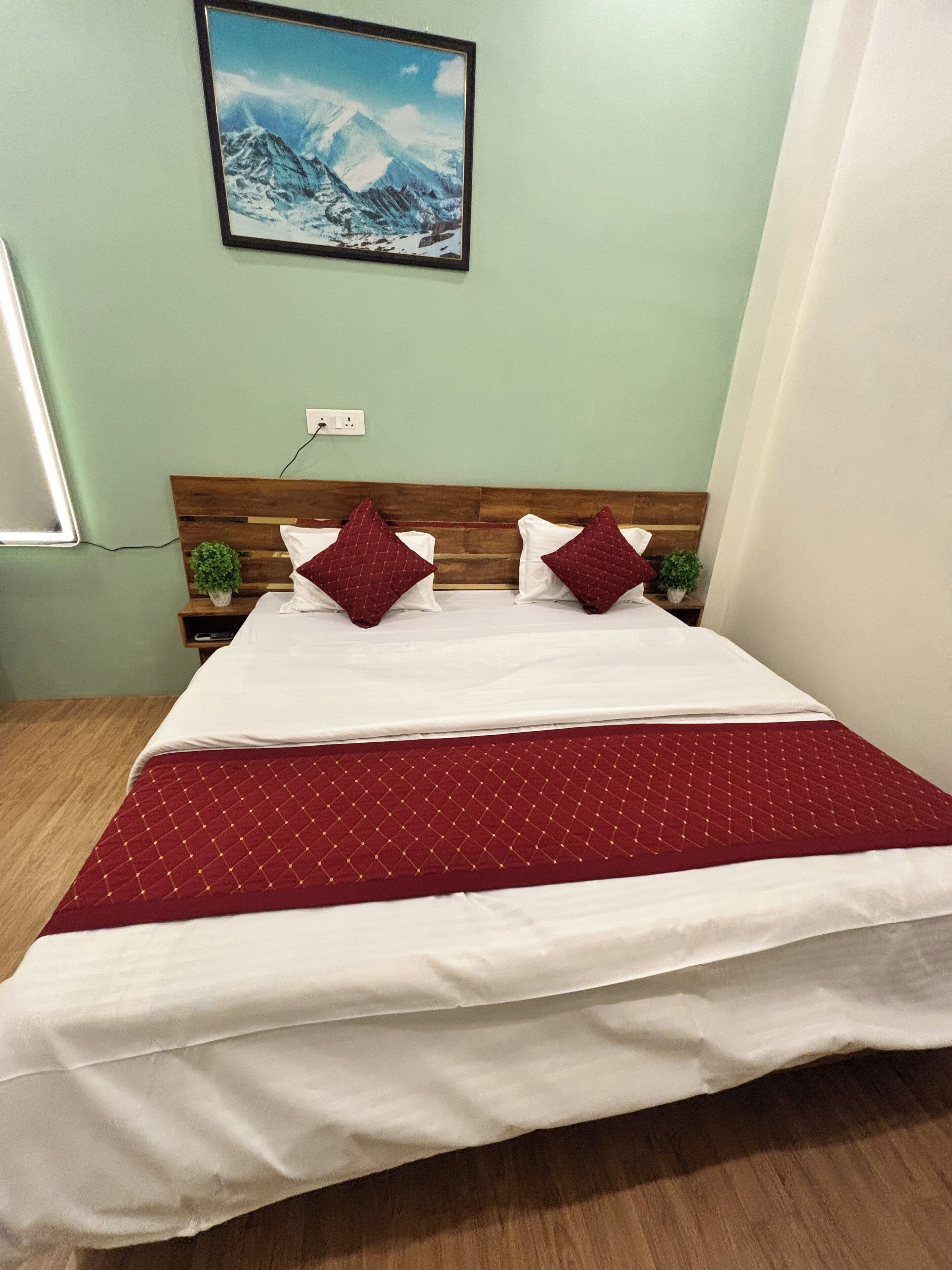 A neatly made bed with a red quilt, two pillows, and a wooden headboard. A small plant is on the nightstand beside the bed.