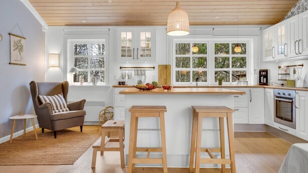 Eine moderne Küche mit weißen Schränken, Holz Arbeitsplatten und einer zentralen Kochinsel mit Sitzgelegenheiten. Fenster bieten Blick ins Grüne, eine Pendelleuchte und eine kleine Sitzecke mit Stuhl und Beistelltisch.