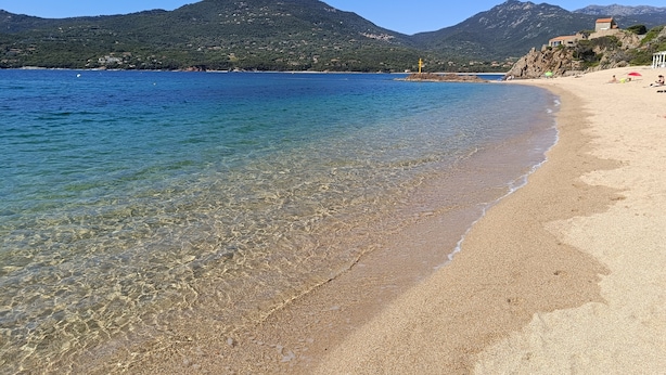 Uma praia com águas claras e rasas, areal arenoso e montanhas ao fundo.