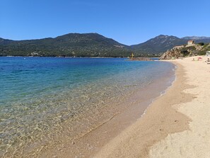 A beach with clear shallow waters, a sandy shore, and mountains in the background.