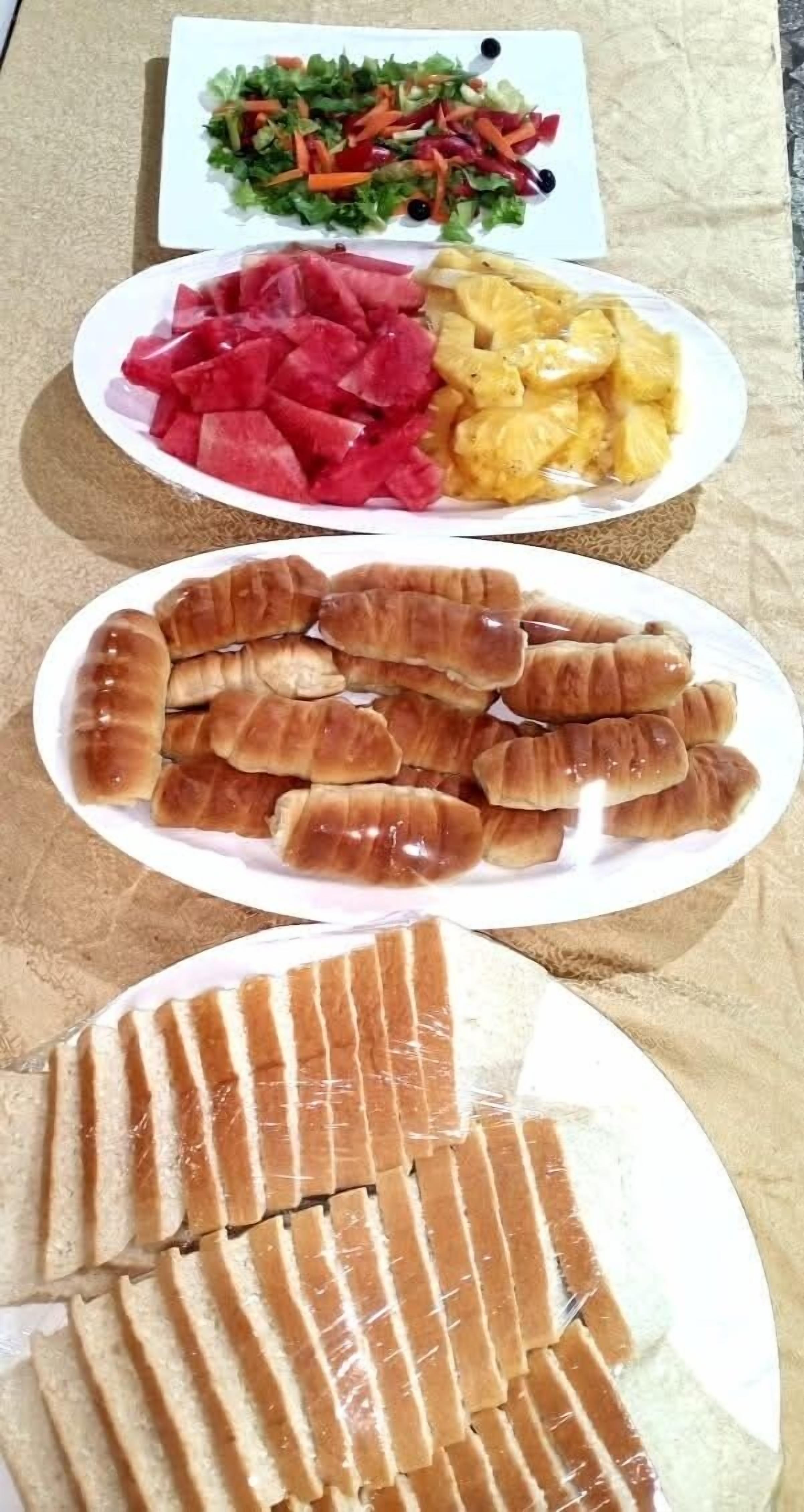 Una mesa con tres platos de comida: una ensalada, sandía y trozos de piña; un plato de cruasanes; y un plato de pan rebanado.