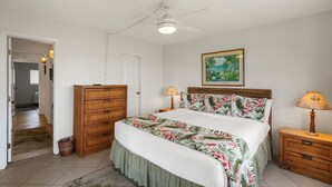 Ein Schlafzimmer mit einem Bett, einem Deckenventilator, einem Nachttisch mit Lampe, einer hölzernen Kommode und einem Bild an der Wand.