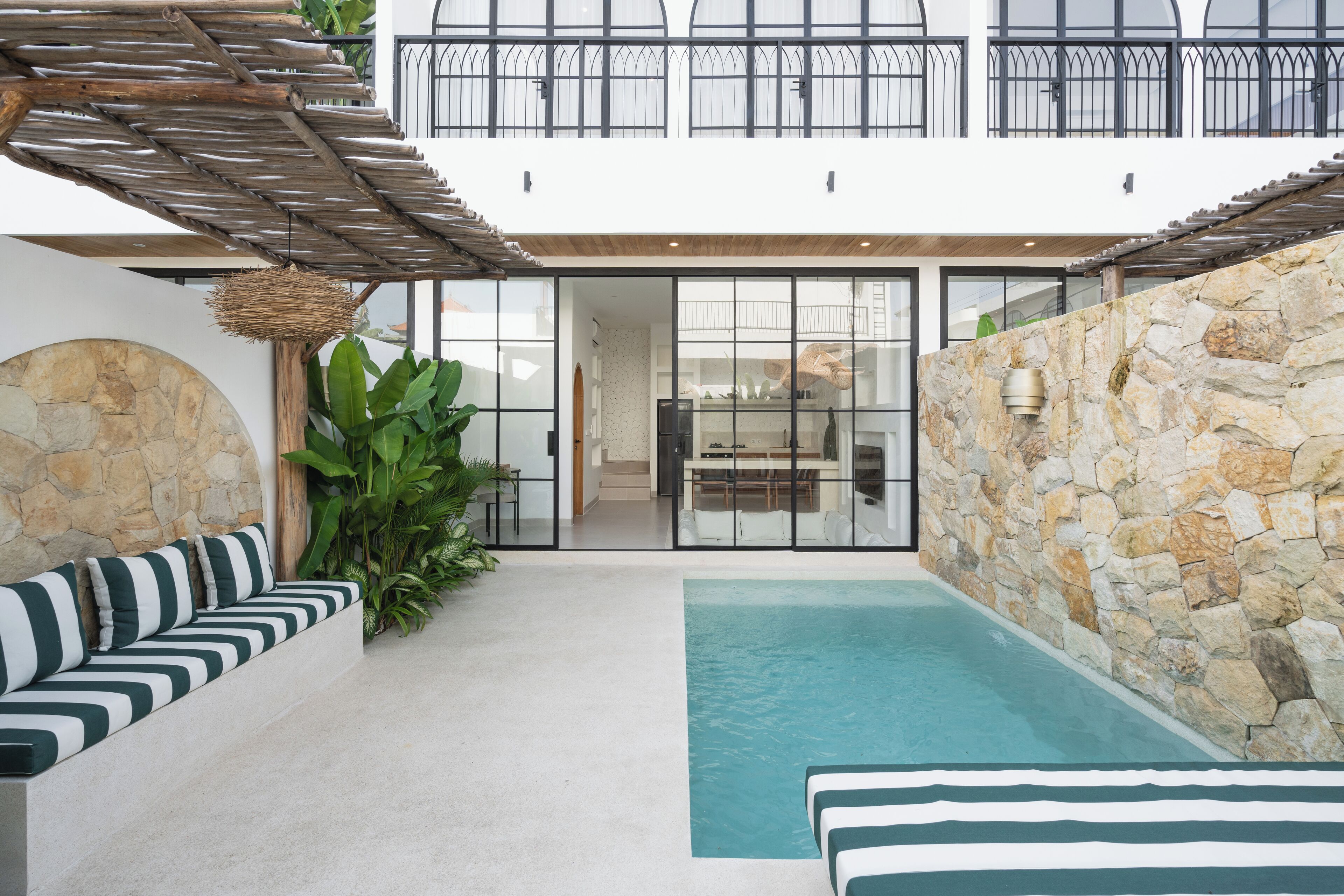 Ein moderner Poolbereich im Freien mit einer Steinwand, einer Sitzecke mit gestreiften Kissen und Blick ins Innere durch Glastüren.
