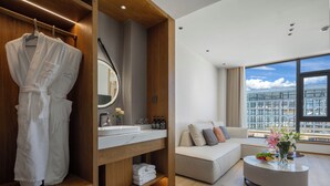 A modern hotel room with a wooden vanity, a large mirror, a sofa, and a view of the cityscape through large windows.