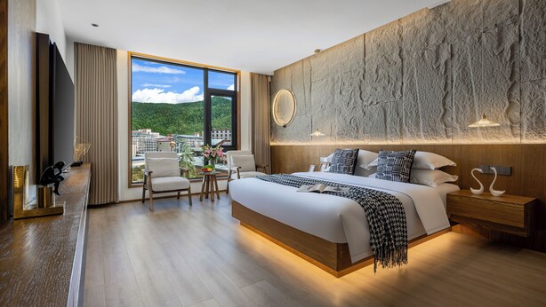 A modern hotel room with a large bed, a wooden headboard, a view of the city, and a small sitting area with a table and chairs.