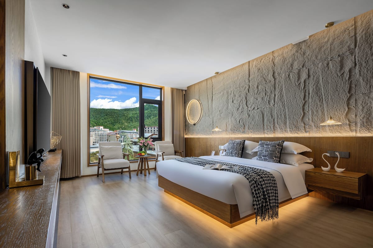 Una habitación de hotel moderna con una cama grande, cabecera de madera, vista a la ciudad y una pequeña zona de estar con una mesa y sillas.