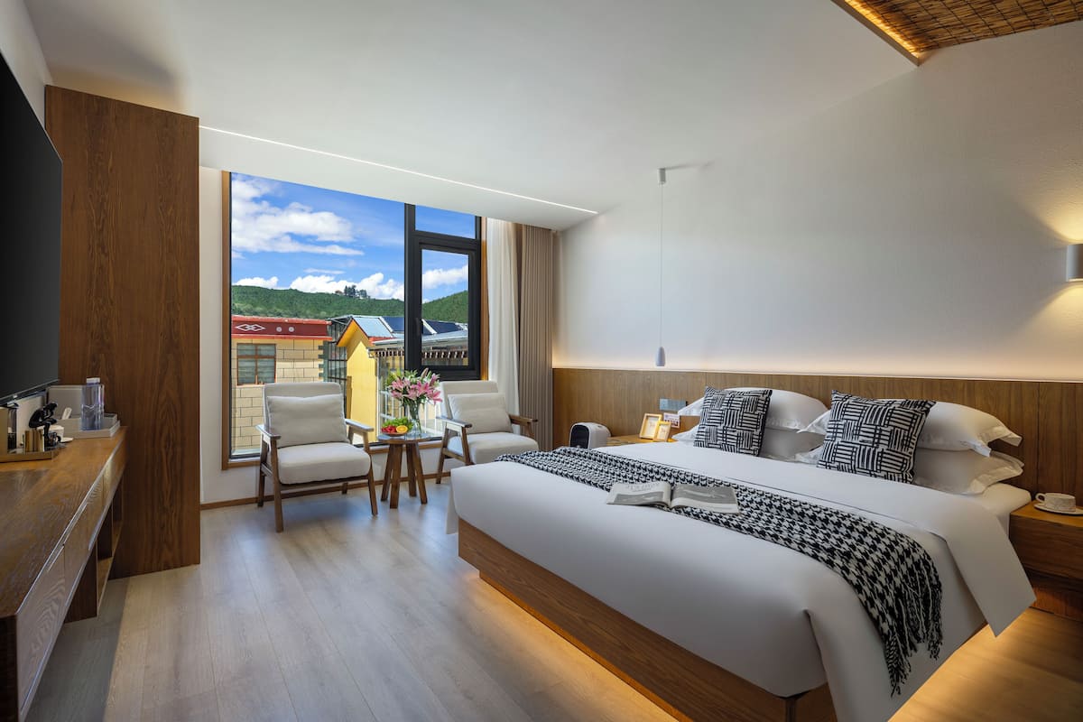 Una habitación de hotel moderna con una cama grande, mesitas de noche, un escritorio y una silla. Se ve una ventana con vistas a edificios y colinas.