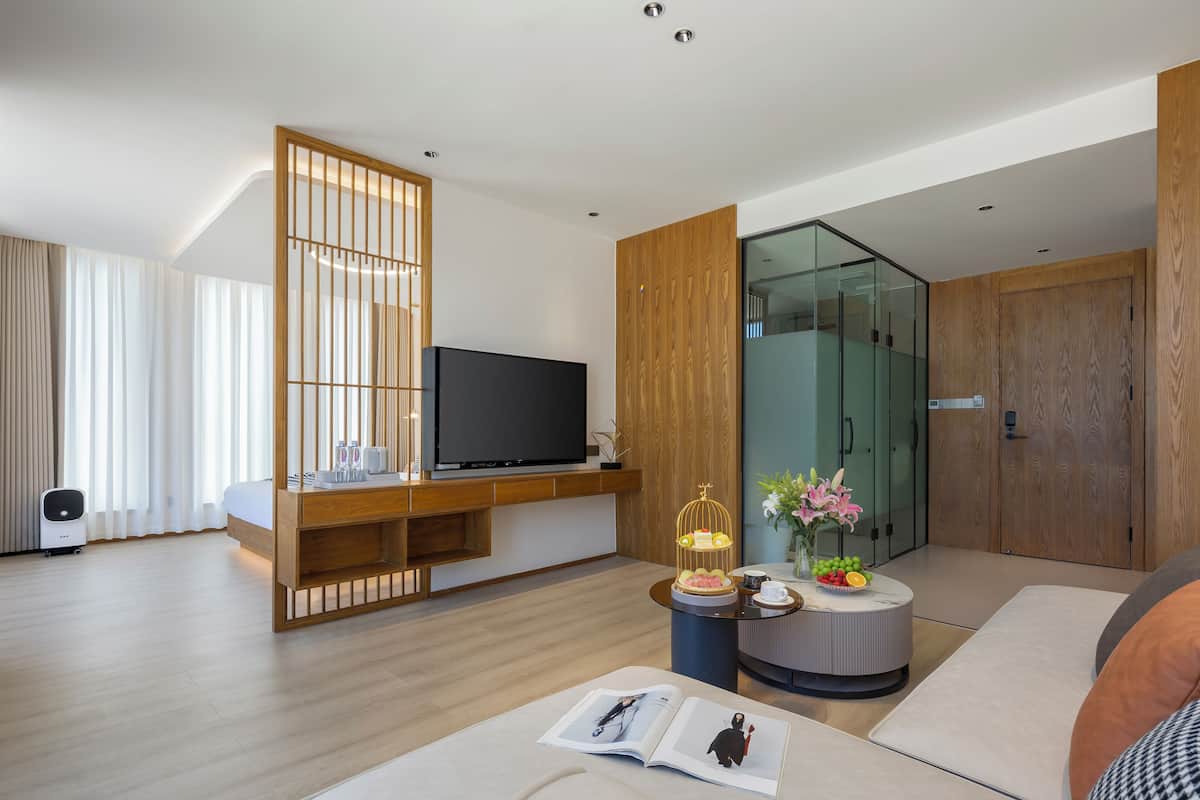 Una habitación de hotel moderna con televisor de pantalla plana, paneles de madera, armario de vidrio, una mesa de centro redonda con un libro y una planta pequeña.