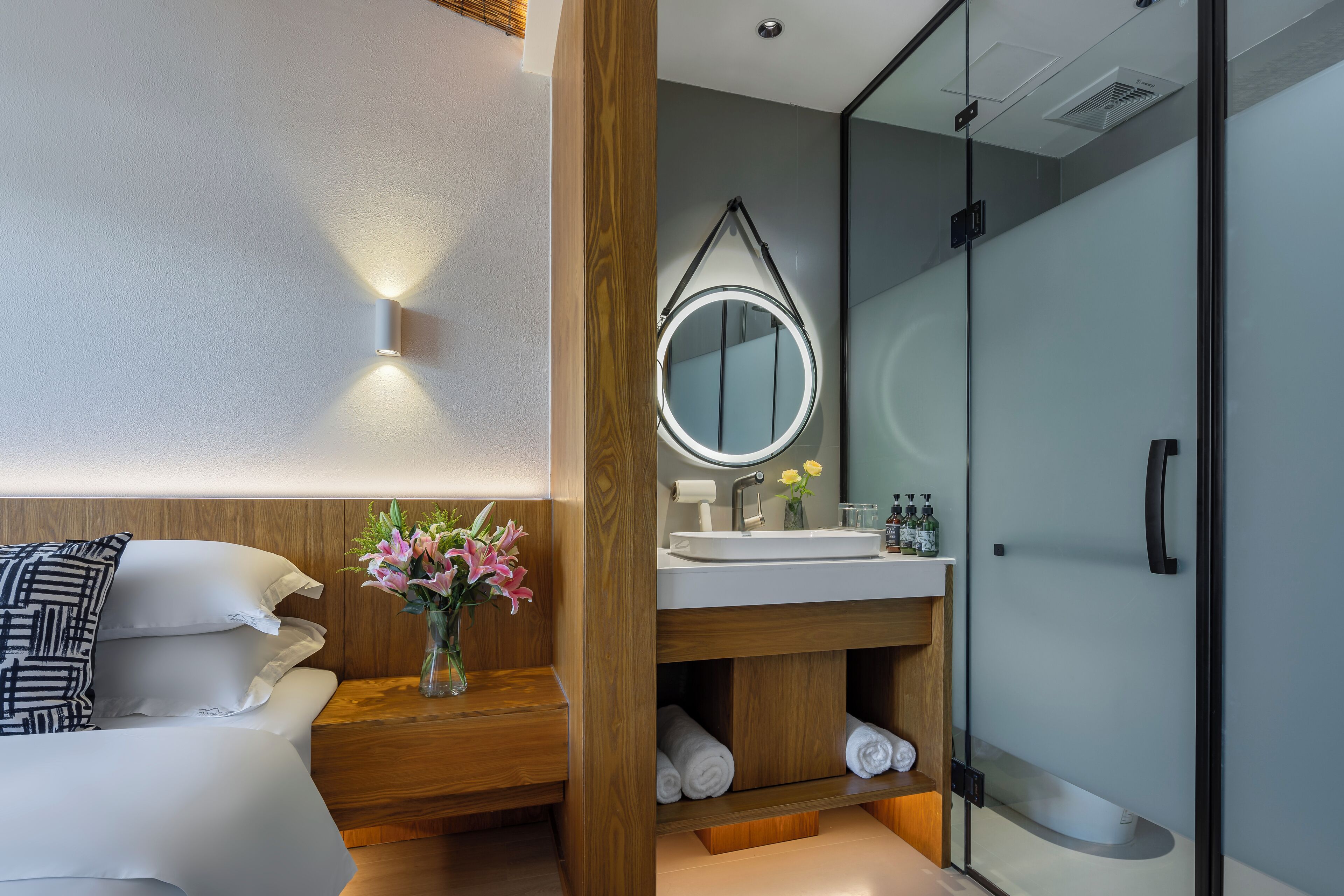 Ein modernes Hotelzimmer mit Bett, Nachttisch, Spiegel, Bad mit Duschkabine aus Glas und Waschtisch aus Holz.