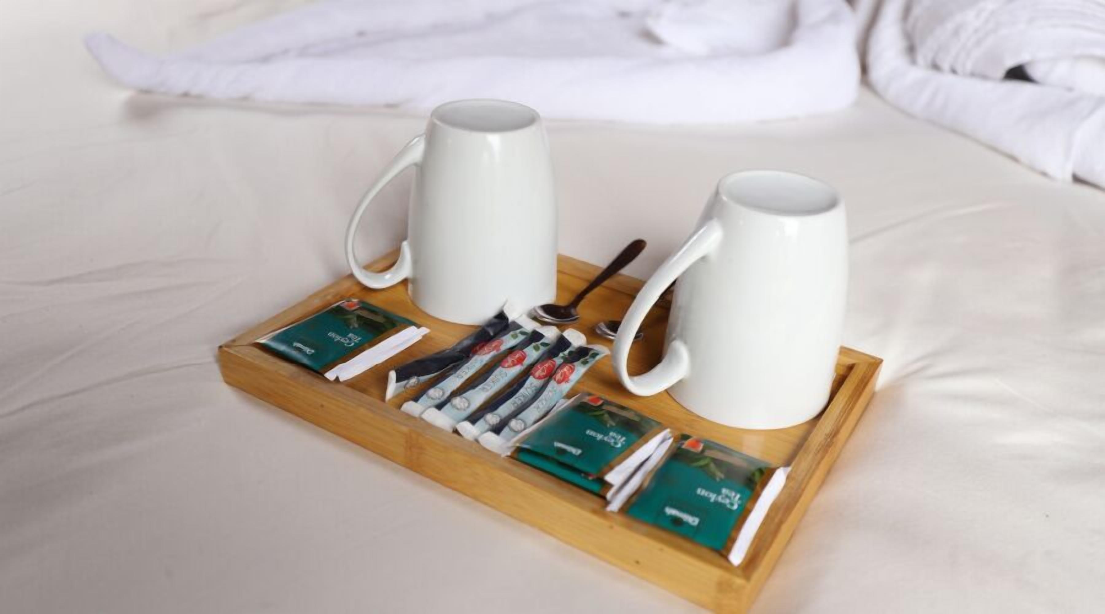 A wooden tray with two white mugs, a spoon, and a set of magazines on a bed.