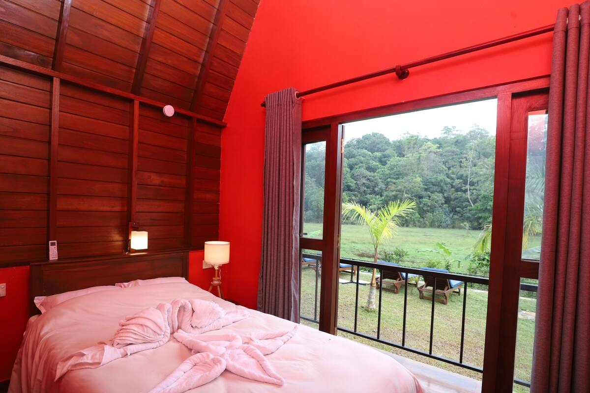 A bedroom with a large bed, a bedside lamp, and a view of a lush garden through a large window.
