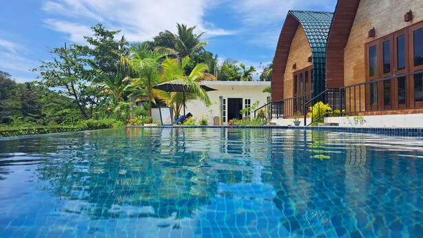 Una piscina cristalina con borde de azulejos, rodeada de abundante vegetación y un edificio con tejado inclinado y amplias ventanas.