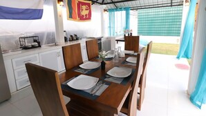 Un comedor con una larga mesa de madera preparada para cuatro, platos blancos y cubiertos. Se observa una zona de cocina con armarios blancos y una bandera colgada en la pared.
