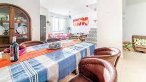 Een eetgedeelte met een tafel gedekt voor vier personen, omringd door leren stoelen. Een houten kast met servies staat links, en in de achtergrond is een kleurrijke wanddecoratie te zien.
