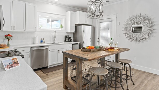 Una cocina moderna con armarios blancos, una isla de madera con taburetes, electrodomésticos de acero inoxidable y un elemento decorativo en la pared.