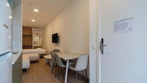 Ein kompaktes Hotelzimmer mit Küchenzeile, einem Bett, einem kleinen Esstisch mit Stühlen und einem an der Wand montierten Flachbildfernseher.