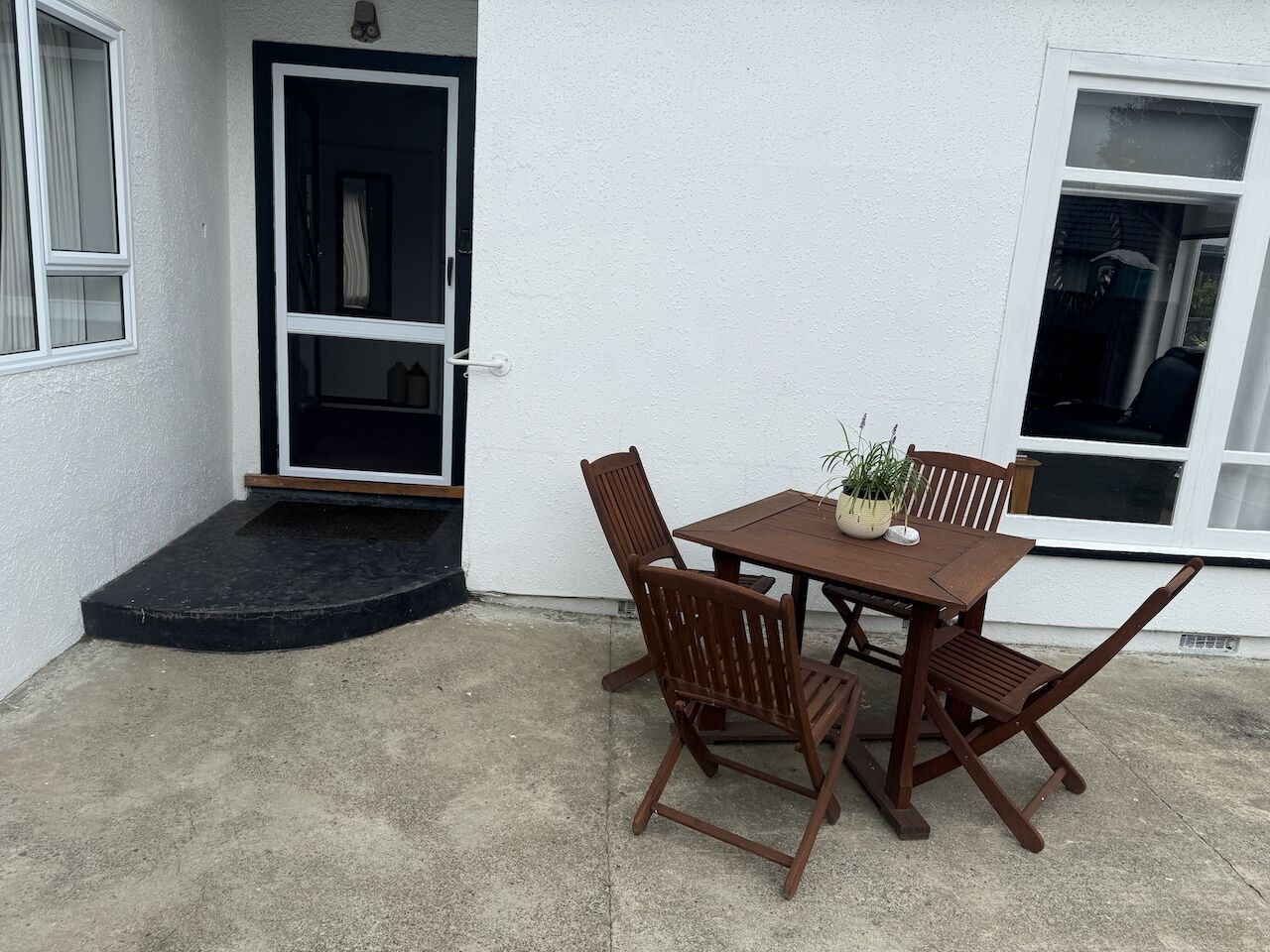 An outdoor patio with a wooden table and chairs, a potted plant, and a door leading to the interior.