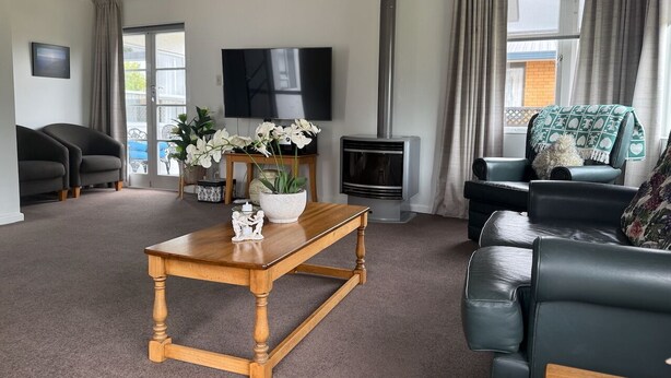 Ein Wohnzimmer mit Couchtisch aus Holz, einer schwarzen Ledersofa, einem Fernseher, einem Kamin und Blick ins Freie durch große Fenster.
