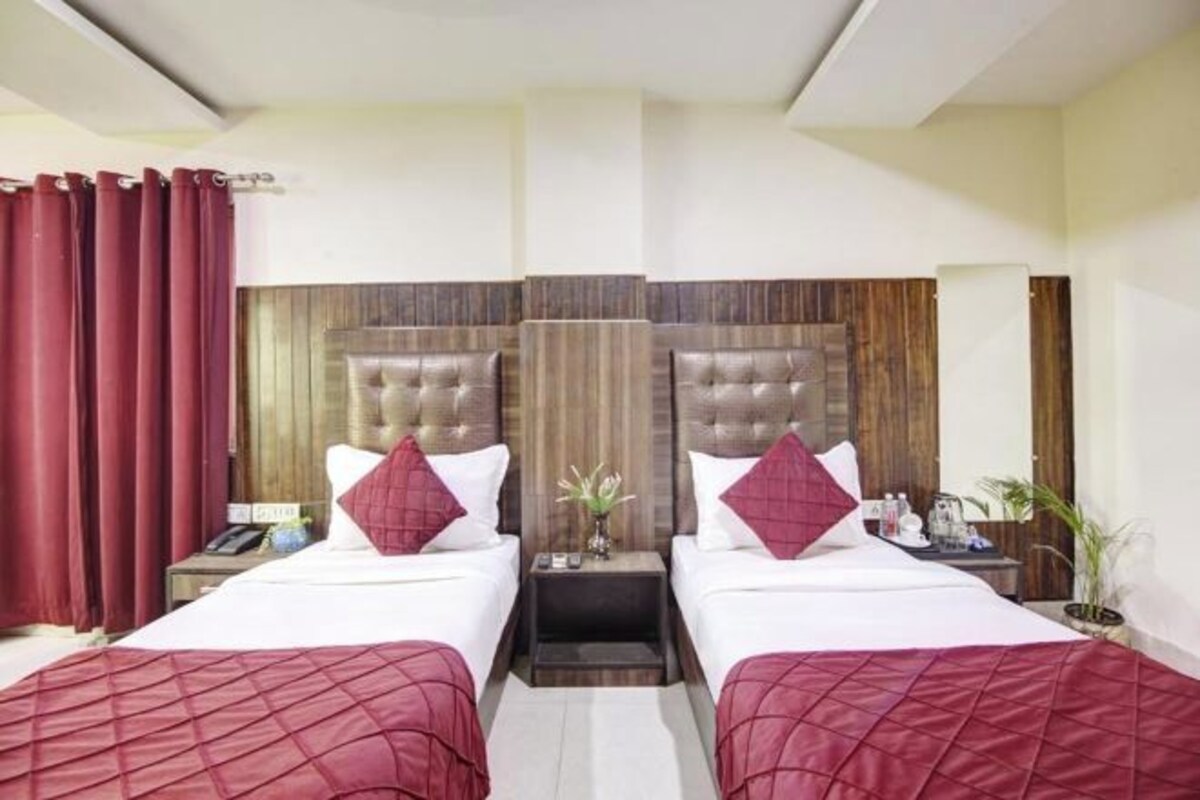 A hotel room with two beds, wooden headboards, red curtains, a bedside table with a lamp, and a small potted plant.