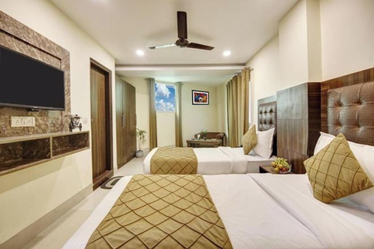 A hotel room with two beds, a flat-screen TV mounted on the wall, a ceiling fan, and a small window with curtains.