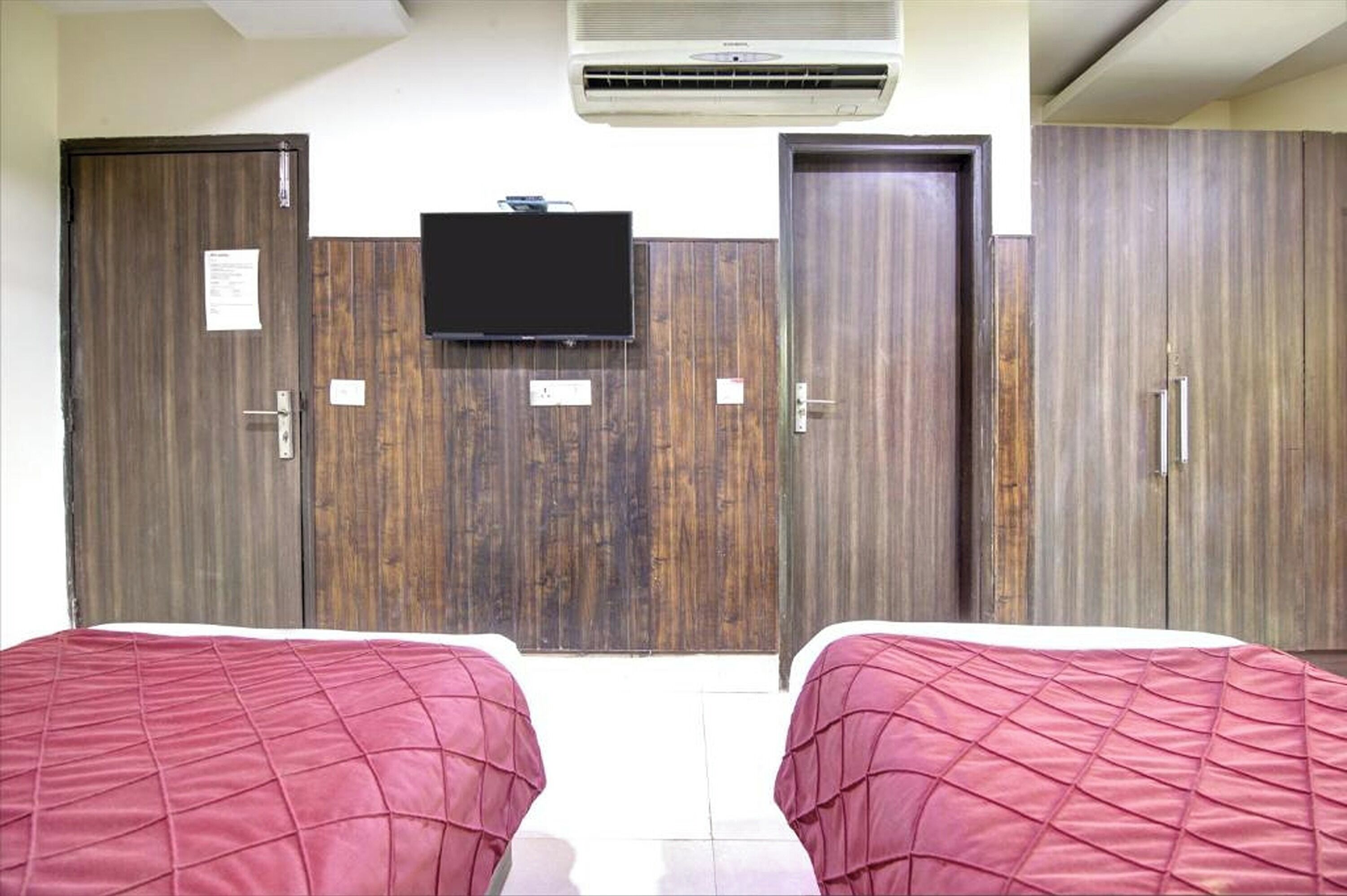 Une chambre avec deux lits, des armoires en bois, un téléviseur à écran plat fixé au mur et un appareil de climatisation placé au-dessus du téléviseur.