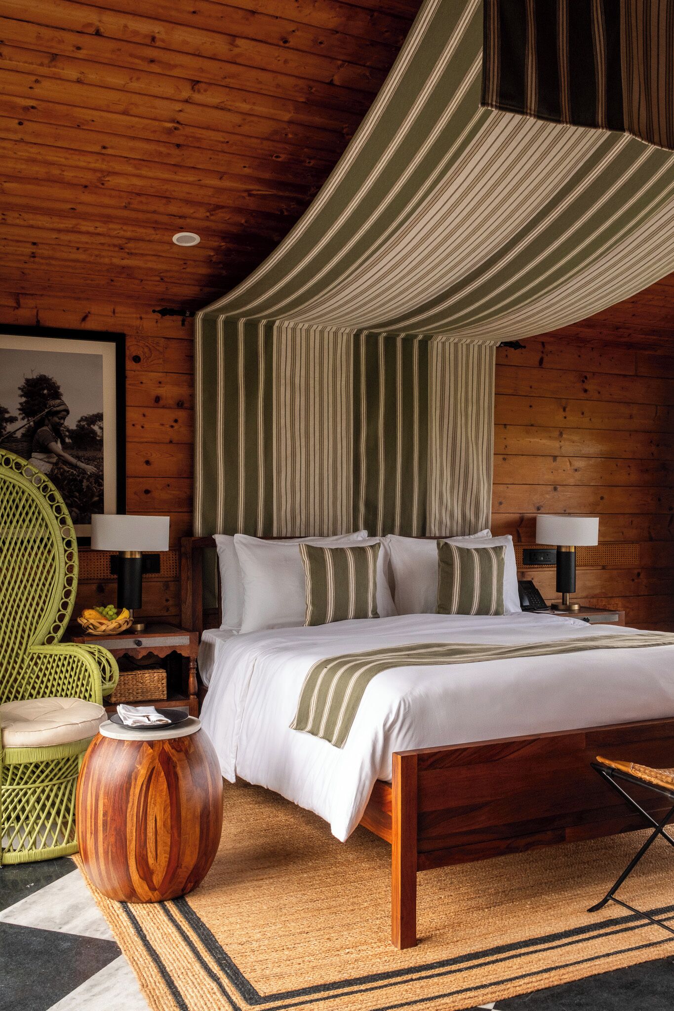 Un dormitorio con paneles de madera, una cama grande, dos mesitas de noche con lámparas, una silla verde y una mesita auxiliar de madera.