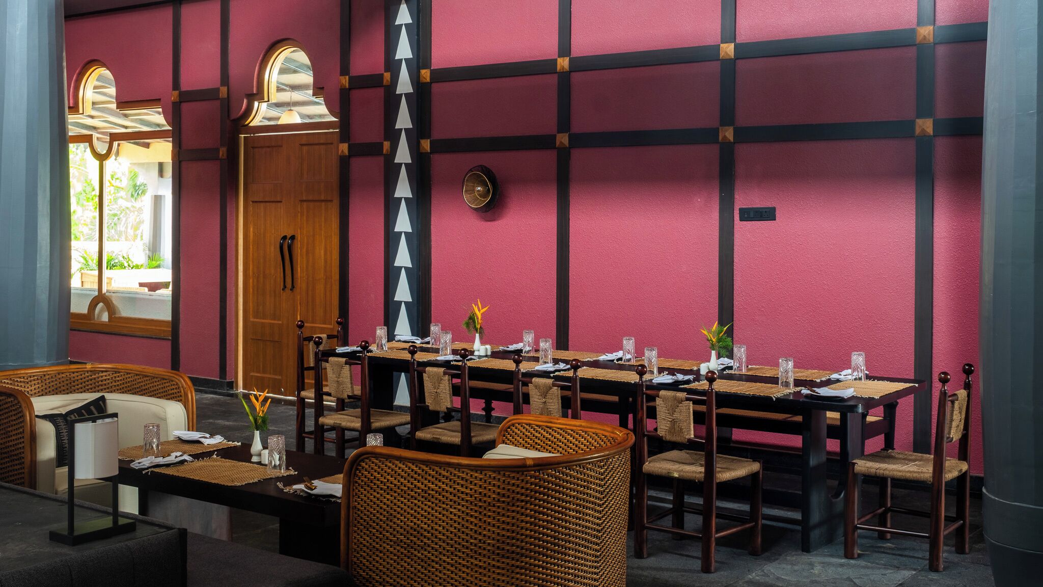 Un comedor con una mesa larga dispuesta para varios comensales, sillas de mimbre y una puerta de madera con ventanal.