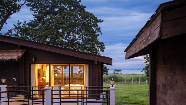 A wooden cabin with a glass door leading to a room, surrounded by a fenced yard and a large tree. - Rhino and River Wildlife Retreat & Spa, Pobitora - Small Luxury Hotels of the World (Mangaldai)