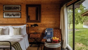 A wooden cabin room with a bed, a desk, a chair, a lamp, and a window with a view of a grassy area. - Rhino and River Wildlife Retreat & Spa, Pobitora - Small Luxury Hotels of the World (Mangaldai)
