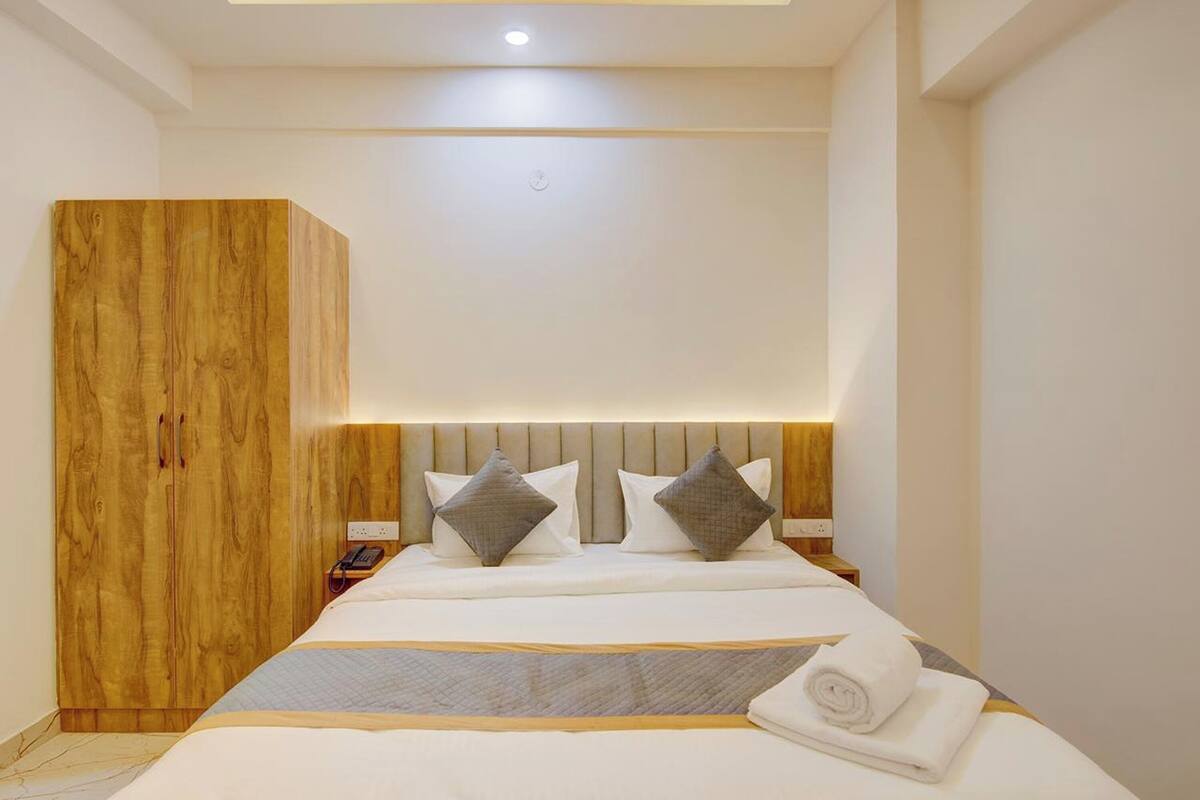 A modern bedroom with a wooden wardrobe, a bed with white bedding and grey pillows, and a bedside table with a lamp.