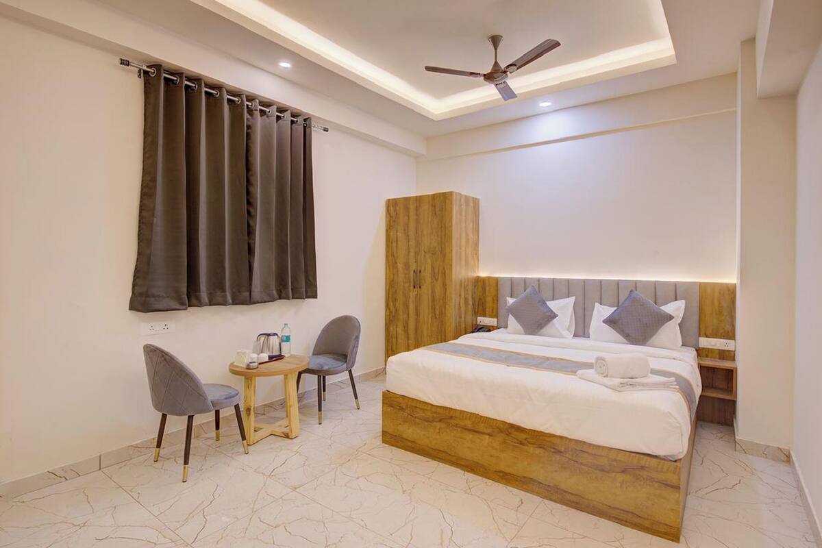 A modern hotel room with a large bed, a desk, two chairs, a ceiling fan, and a wooden wardrobe.