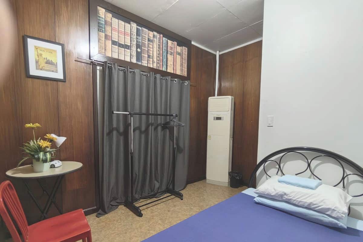 Una stanza con un letto, una sedia, un tavolino con un vaso, una tenda e una mensola a parete con libri.