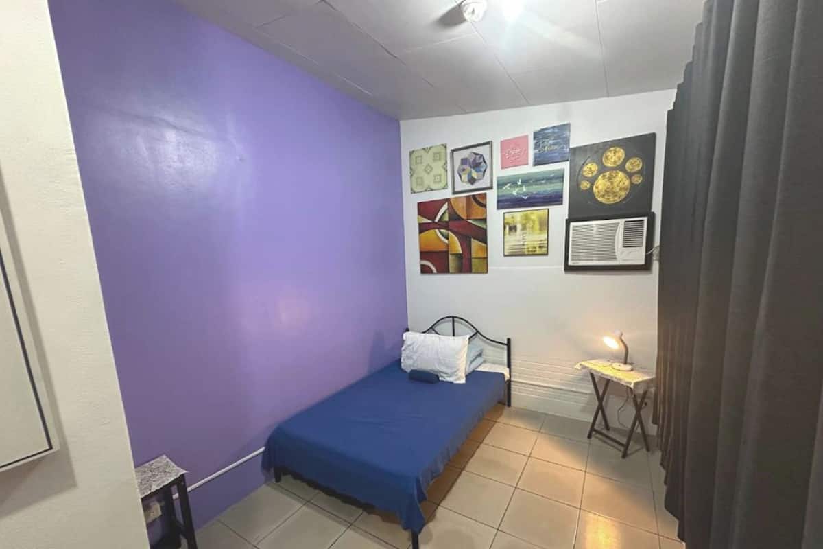 Una stanza piccola con un letto singolo, un tavolino e una parete decorata con diverse opere d'arte.
