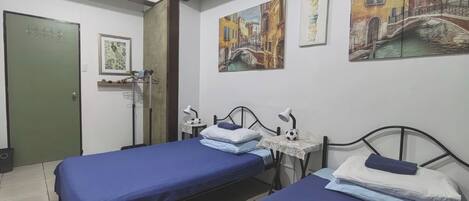 A hotel room with two beds, a desk with a lamp, a chair, a green door, and artwork on the walls.