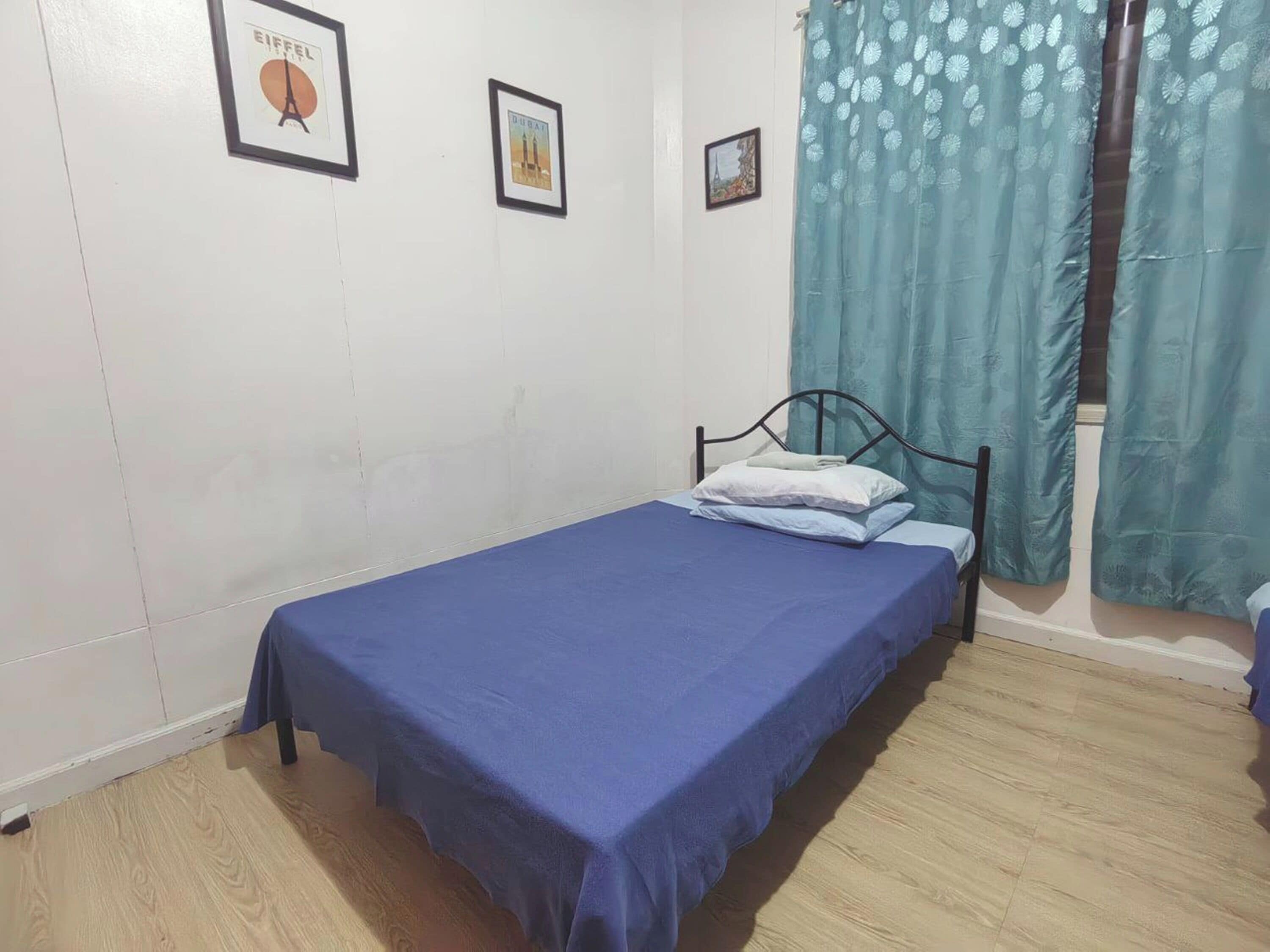 Una habitación pequeña con una cama individual, colcha azul, estructura de cama de metal y cortina color turquesa.