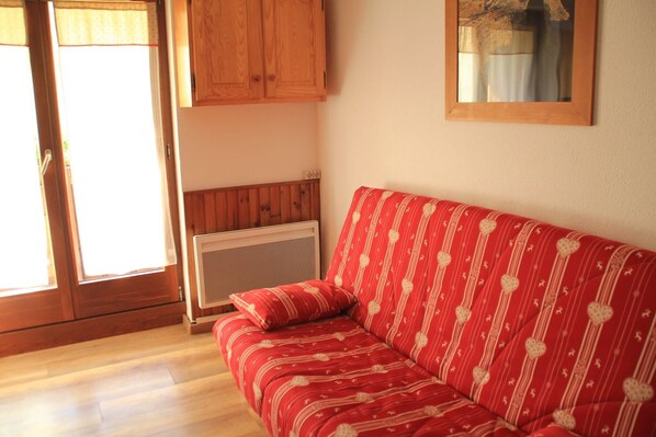 A red sofa with heart patterns, a wooden cabinet, and a radiator in a room with wooden flooring and a window.