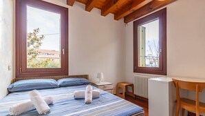 Un dormitorio con cama, dos ventanas, un radiador, un escritorio y una silla.