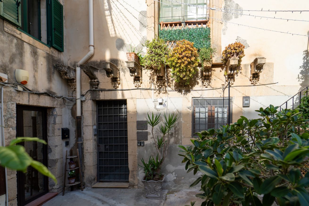 A narrow alleyway with a building featuring a barred door, a small window with a grid pattern, and hanging plants.