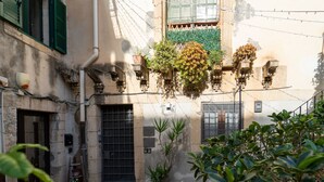 Une ruelle étroite bordée d’un immeuble dont la porte est munie de barreaux, avec une petite fenêtre à carreaux et des plantes suspendues.