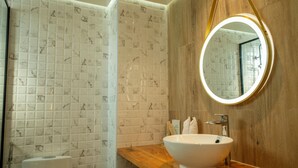 A bathroom with a white marble tiled wall, a wooden vanity, a round mirror, and a white sink. - Athiri Residence Dhiffushi (Dhiffushi)