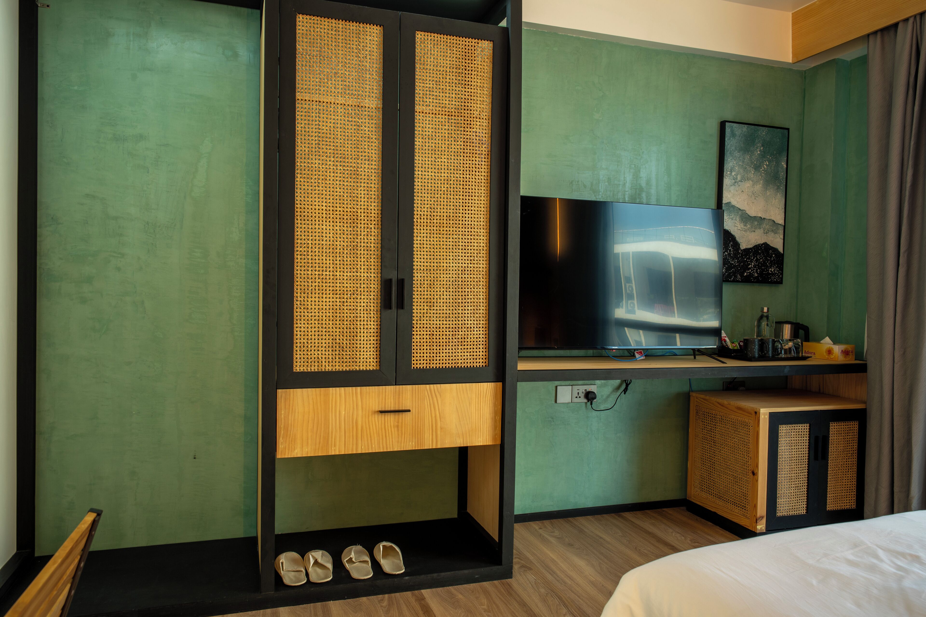 Una habitación de hotel con una cama, un televisor, un armario de madera con zapatos y una pared verde.