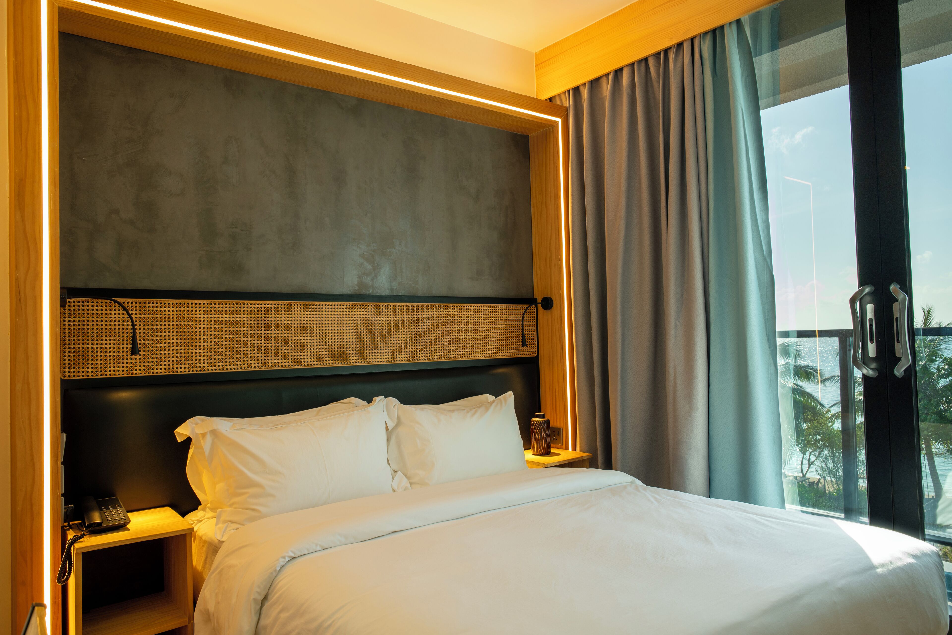 Habitación de hotel con una cama grande, una mesita de noche con lámpara, un teléfono y vistas al exterior a través de una ventana.