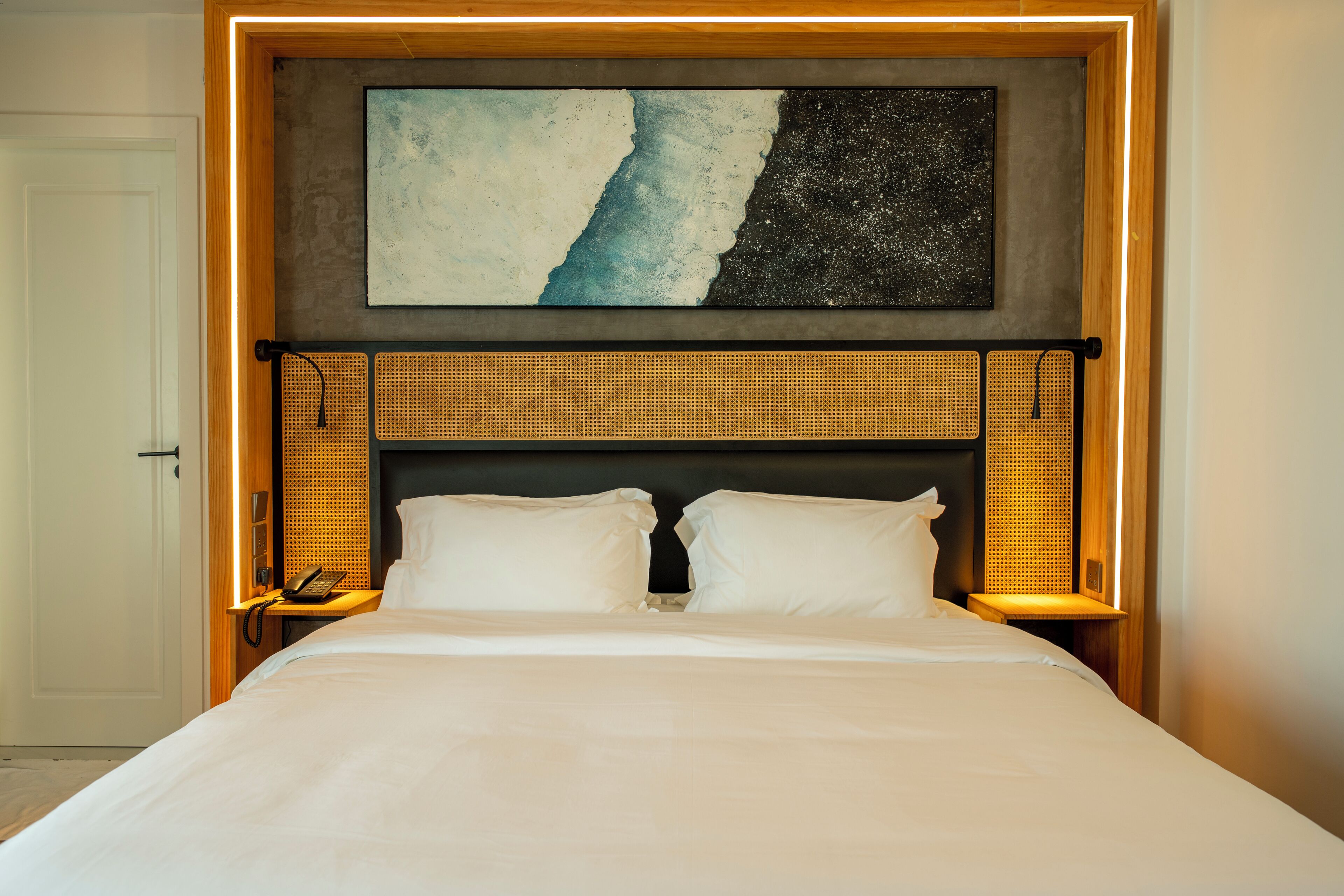 Una cama bien hecha con un cabecero de diseño trenzado, una obra de arte colgada en la pared con un diseño abstracto en azul y negro, y un teléfono en la mesita de noche.