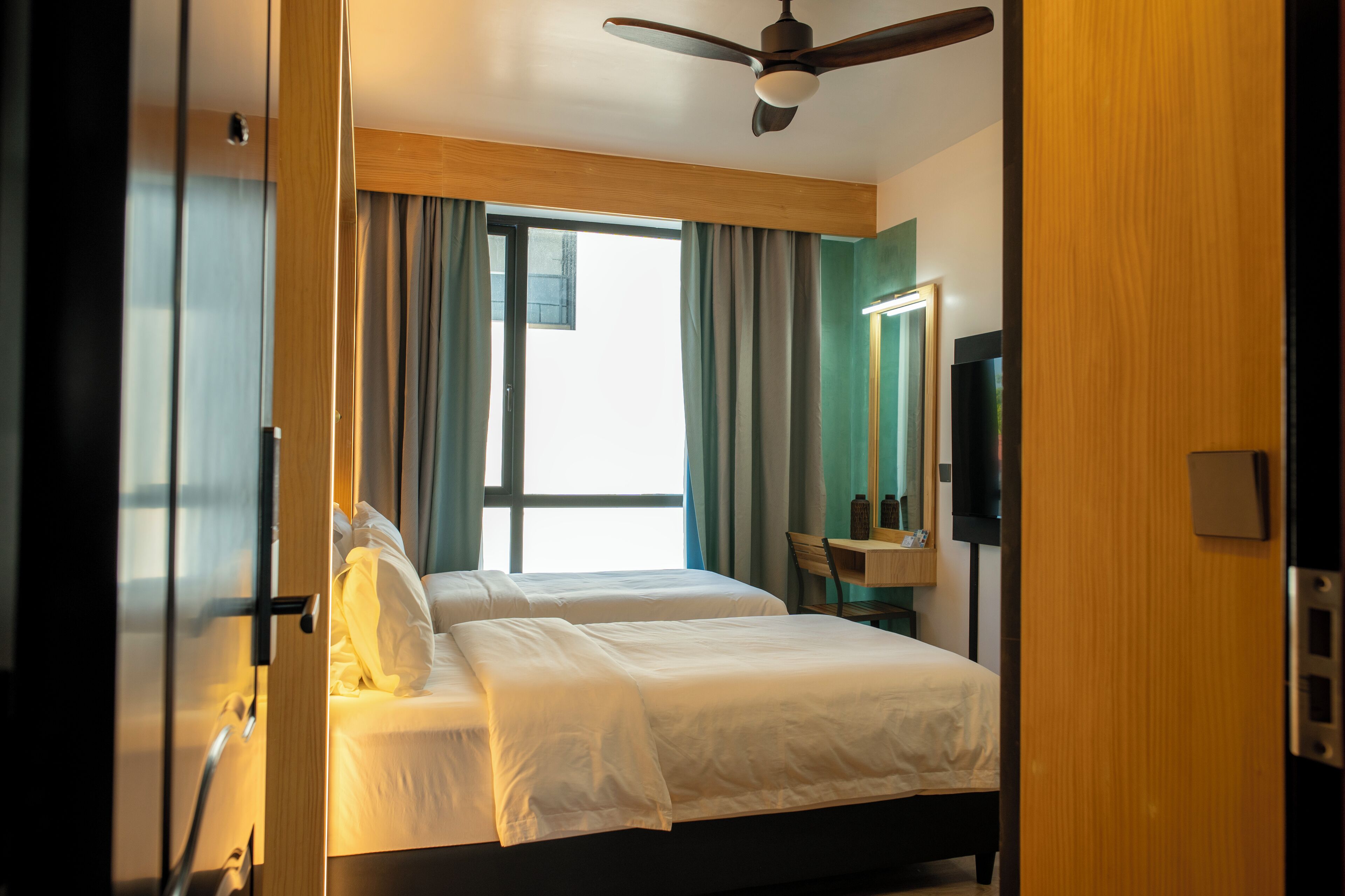 A hotel room with a bed, a ceiling fan, a desk with a chair, a mirror, and a window with curtains.