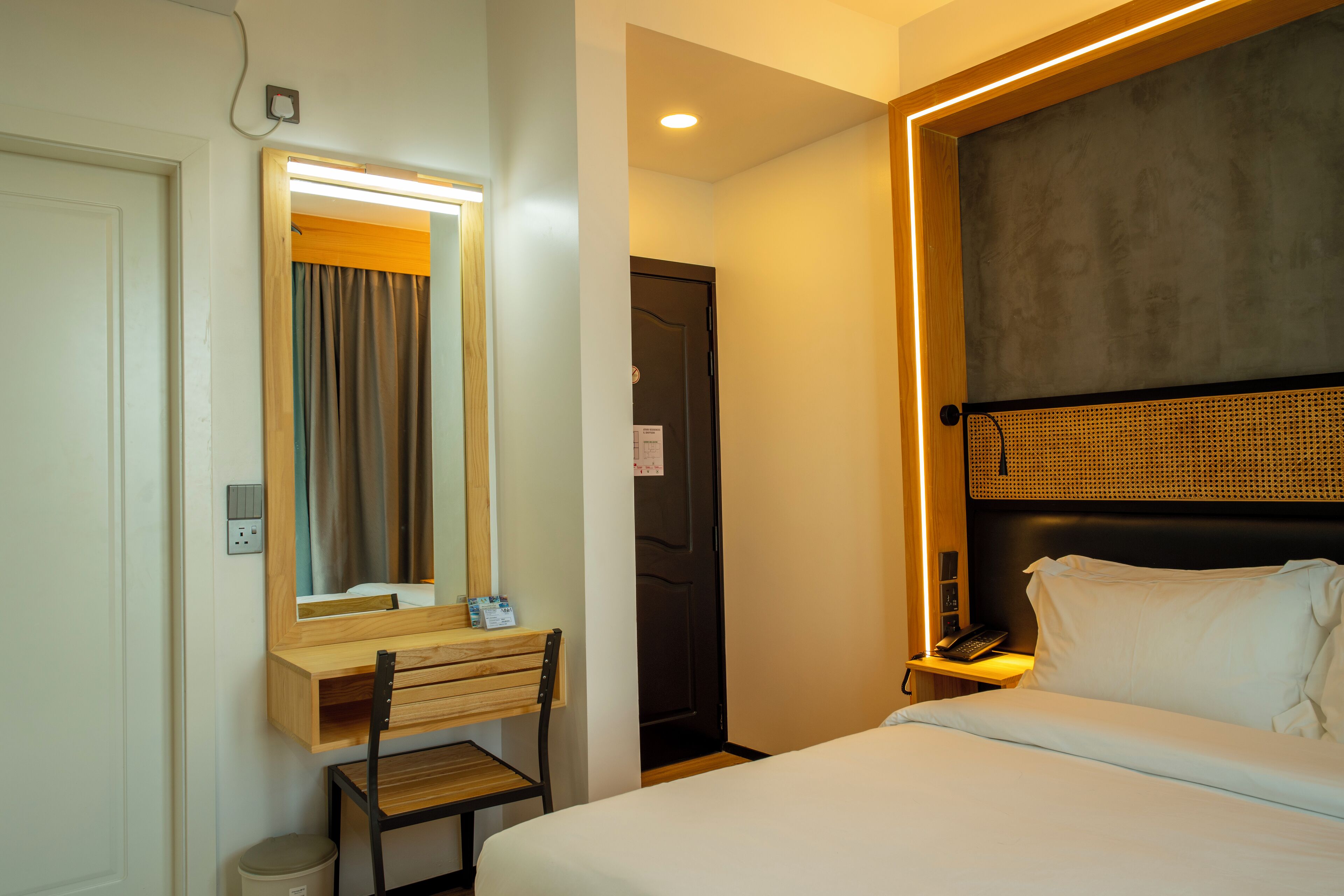 A hotel room with a bed, a desk with a chair, a mirror, a bedside table with a lamp, and a door.