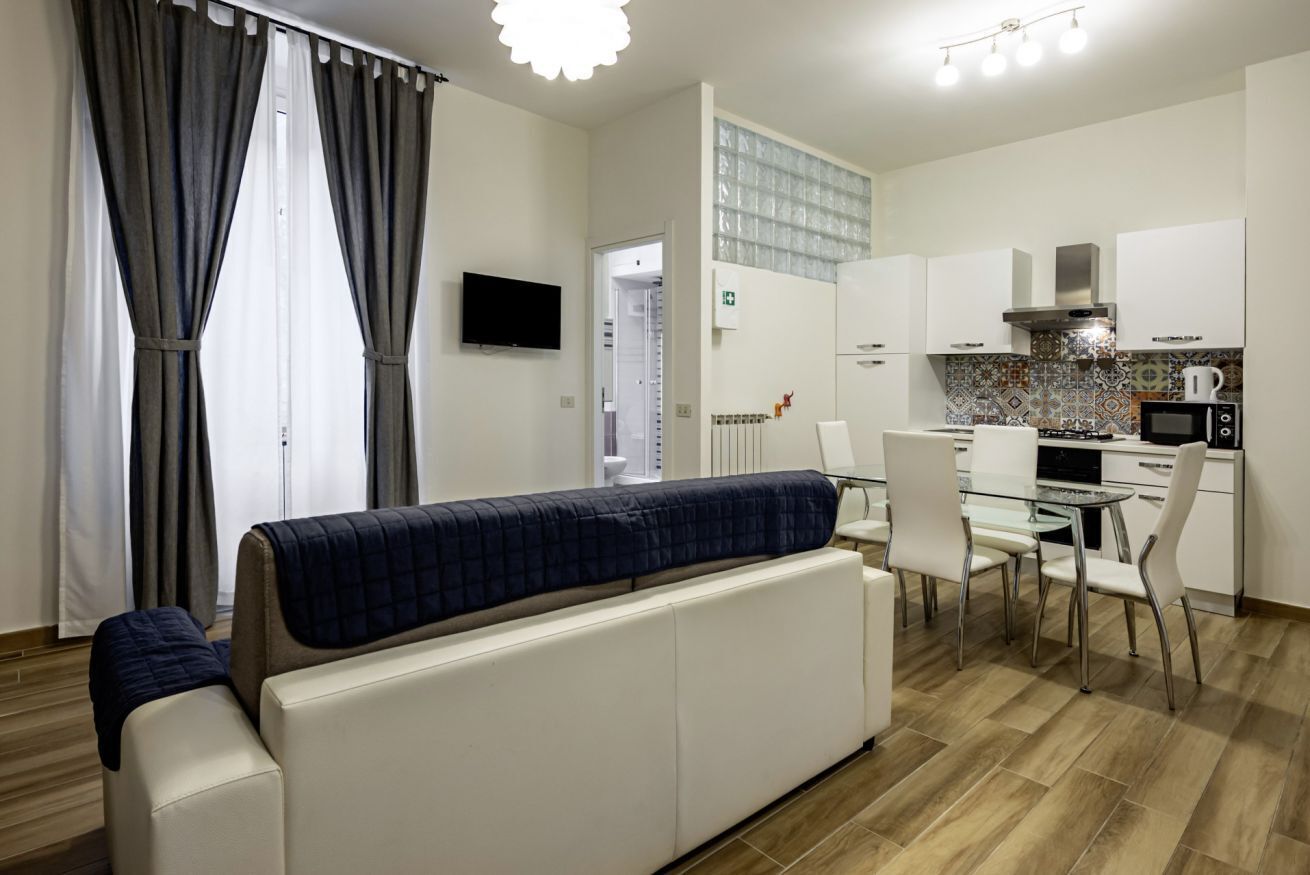 Uma sala moderna com sofá, mesa de jantar com cadeiras, cozinha com armários e eletrodomésticos, e uma TV fixada na parede.