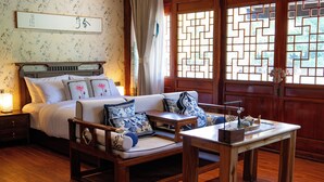 A traditional-style bedroom with a wooden bed, a sofa with blue and white cushions, a wooden table, and a window with a geometric pattern. - Long Chuan River Kwai (Thong Pha Phum)