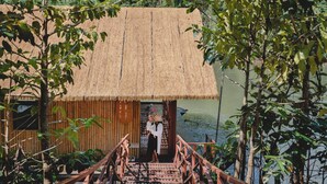 A wooden structure with a thatched roof, a wooden staircase, and a view of a body of water. - Long Chuan River Kwai (Thong Pha Phum)