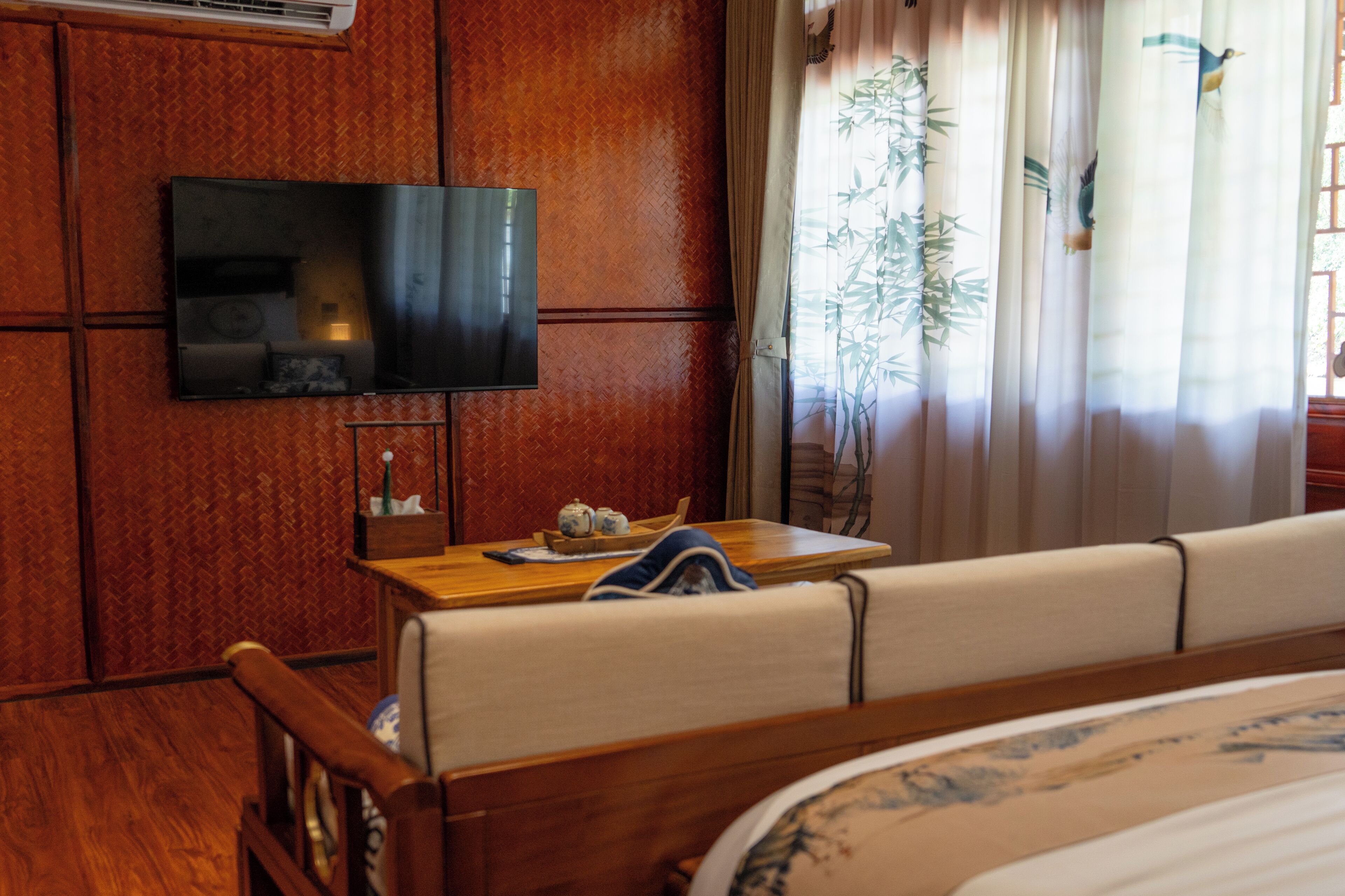 Une chambre d’hôtel avec un mur en bois, un téléviseur à écran plat fixé au-dessus d’une table en bois, un lit recouvert d’une courtepointe à motifs floraux et une fenêtre ornée de rideaux en bambou.